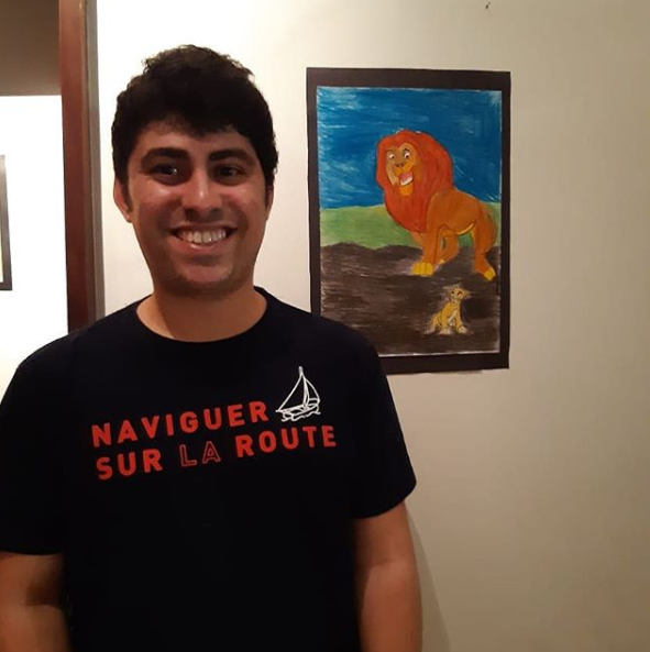 #pracegover: representa a foto de um jovem rapaz sorrindo , e na parede ao fundo encontra-se um quadro que ele produziu